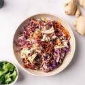 A colorful salad with shredded purple cabbage, carrots, and dressing in a beige bowl on a white surface. Fresh cilantro and ginger root surround it, suggesting fresh ingredients.
