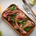 Carne asada marinated in citrus juices sliced thin on a plate with cilantro, limes, and orange slices.
