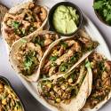 A platter of chicken tacos topped with cilantro and corn, accompanied by a bowl of green sauce, on a light background.