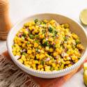 A vibrant corn salad with chopped red onions and cilantro in a white bowl, set on a fringed orange cloth. Lime slices and a pepper grinder nearby.