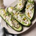 Four cucumber “boats” filled with creamy shredded chicken salad, garnished with chopped pickles, sliced green onions, and a sprinkle of dried dill, arranged on a white oval plate with a light blue cloth napkin nearby.
