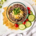 Easy black bean dip in a bowl with shredded cheese and cilantro with cucumbers, tomatoes, and chips on the side.