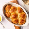 Golden brown dinner rolls in an oval dish on a wooden board. Nearby, a small bowl of melted butter with a spoon, and two forks on stacked plates. Warm, inviting tone.