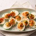A plate of deviled eggs garnished with paprika and chives is set on a beige napkin. A small bowl of chives and a pepper mill are nearby.