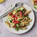 A vibrant plate of pasta mixed with cherry tomatoes, broccoli, and herbs, accompanied by a glass of olive oil and fresh basil leaves.