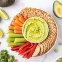 Edamame hummus with frozen edamame, cilantro, avocado, and lime on a plate with vegetables and crackers.