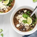 Black bean soup in two bowls with black beans, tomatoes, and onions topped with cilantro, limes, and cheese.