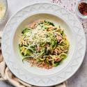 Garlic Parmesan zucchini noodles served in a white dish, topped with grated cheese and red pepper flakes, accompanied by ingredients like olive oil and shredded Parmesan cheese.