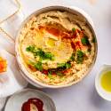 Hummus made with chickpeas in a bowl with olive oil, parsley, and paprika with carrots and pretzels on the side.