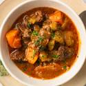 A bowl of hearty beef stew with chunks of carrot, celery, and garnished with fresh parsley, served on a cream-colored plate.
