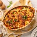 Baked lasagna in an oval dish on a checkered towel, garnished with chopped parsley. A pepper grinder and a plate with utensils are nearby.