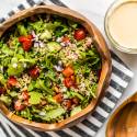 Mediterranean quinoa bowl with quinoa, arugula, red peppers, tomatoes, and hummus dressing in a wooden bowl.
