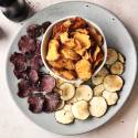 A platter of assorted vegetable chips in three colors: purple, golden, and white, served alongside a small bowl of salt.