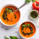 Roasted tomato basil soup in two bowls with pesto and fresh basil leaves.