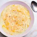 Slow cooker cheesy cauliflower soup with a metal spoon, shredded cheese, and black pepper.