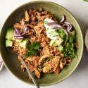 A chicken burrito bowl topped with avocado, cilantro, red onions, sour cream, and chopped chives, served in a green ceramic dish with a spoon.