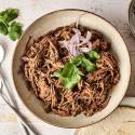 A bowl of tender, shredded Mexican beef adobo is garnished with fresh cilantro and sliced red onions, ready to be served with tortillas.