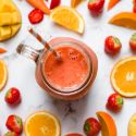 Strawberry mango smoothie in a glass with fresh strawberries, mangoes, and carrots on the side.