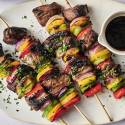 Grilled kebabs featuring marinated beef and colorful vegetables, served with a small bowl of dipping sauce on a white plate.