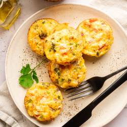 Plate of five colorful egg muffins with veggies, garnished with a parsley leaf. Accompanied by a black fork, napkin, and nearby olive oil. Inviting and fresh.