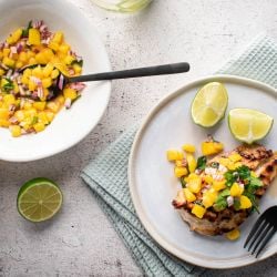 Grilled chicken with mango salsa on a plate with fresh limes and a bowl of mango salsa on the side.