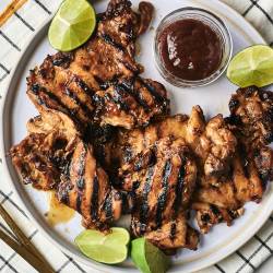 Grilled chicken thighs garnished with lime wedges served on a plate with a side of barbecue sauce. Elegant utensils nearby.