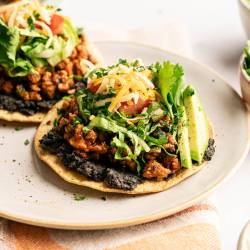 Loaded ground turkey tostadas topped with black beans, seasoned turkey, shredded lettuce, diced tomatoes, shredded cheese, fresh cilantro, and avocado slices, served on a plate with extra toppings nearby.