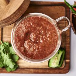 A glass jar filled with chunky salsa rests on a wooden board, surrounded by fresh cilantro and jalapeños, with vibrant chili peppers nearby.