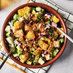 A colorful salad in a brown bowl, featuring diced fruits, veggies, and spices, served with metal spoons on a checkered towel.