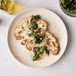 Overhead view of roasted cauliflower steaks garnished with chimichurri sauce, served on a plate with olive oil and pepper mill nearby.