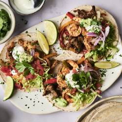 Three vibrant tacos topped with shrimp, beef, lettuce, tomatoes, onions, and cilantro, served with lime wedges and dipping sauce.