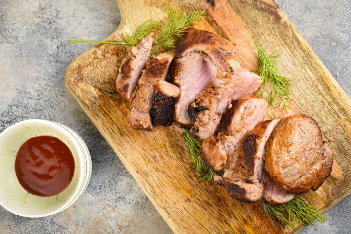 Barbecue rubbed pork tenderloin sliced on a wooden board with rosemary.