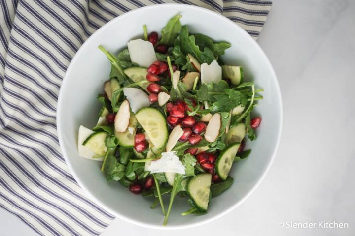 Arugula, cucumber, and pomegranate salad in a bowl with shaved Parmesan cheese.