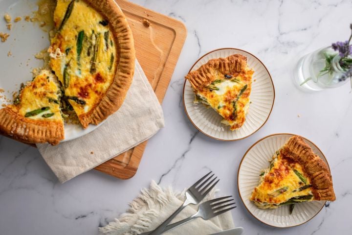 Asparagus quiche with eggs, shredded cheese, asparagus, and red onions cooked in a golden brown crust and served on plates.