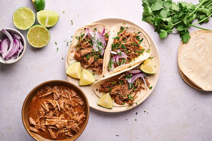 Slow cooker beef barbacoa tacos on corn tortillas with red onion, cilantro, and lime wedges.