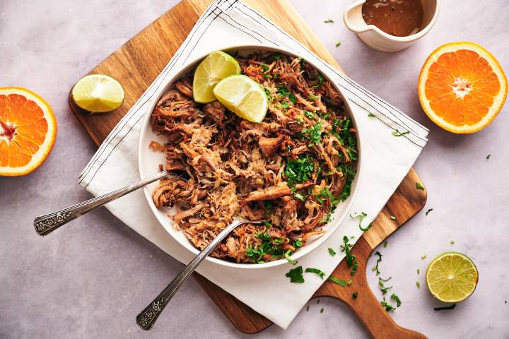 Carnitas served with shredded pork in a bowl with lime wedges, oranges, and cilantro.