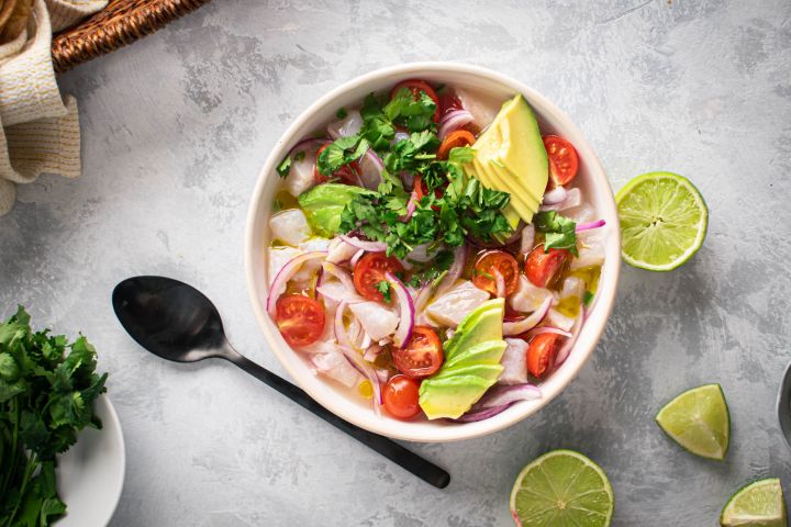 Ceviche with fish marinated in lime juice and served with cilantro, avocado, tomatoes, and red onion in a white bowl.