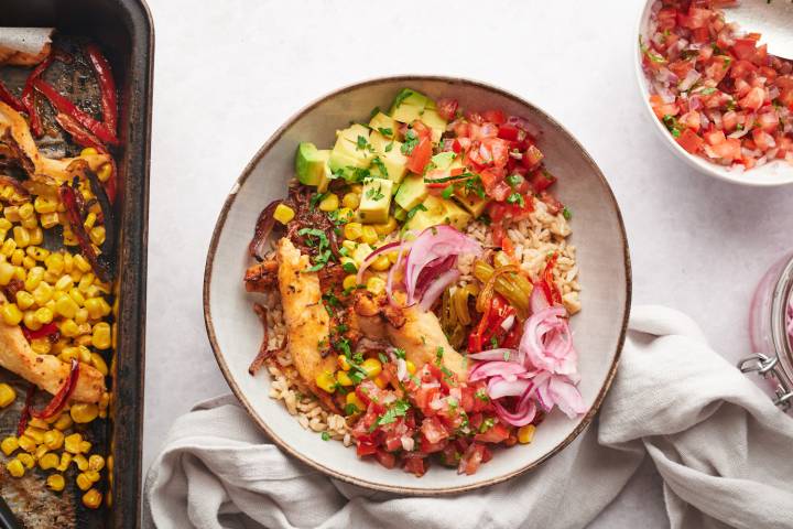 Chicken fajita bowls with seasoned chicken, peppers, onions, corn, and salsa in a bowl with a sheet pan on the side. 