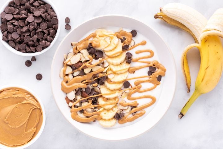 Chunky monkey banana peanut butter Greek yogurt bowl with sliced bananas, almonds, chocolate chips, and melted peanut butter.