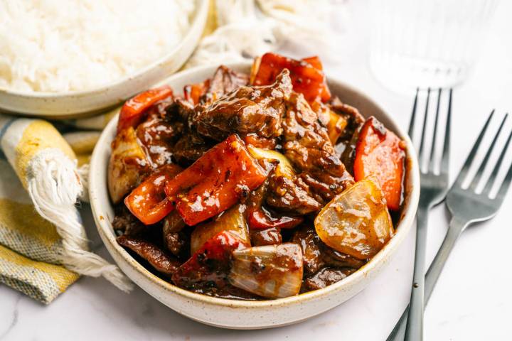 A bowl of beef stir-fry with red peppers and onions in a savory sauce on a table next to a plate of white rice, a napkin, and forks.