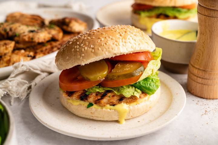 A grilled chicken burger with lettuce, tomato, pickles, and mustard in a sesame bun on a plate. Grilled chicken and sauce in the background.