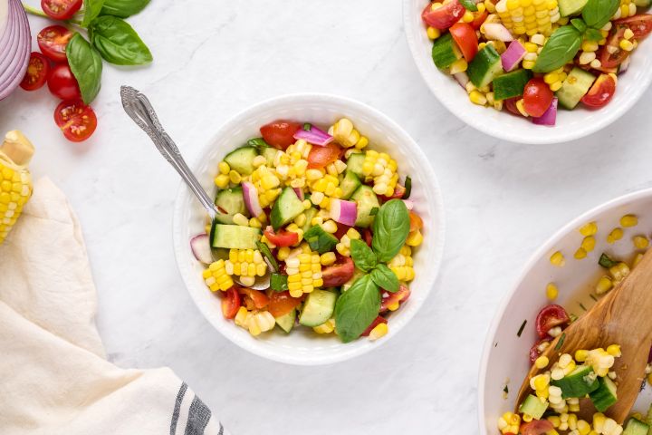 Corm salad with fresh corn, cucumbers, cherry tomatoes, and red onion in a bowl with vinaigrette and basil.