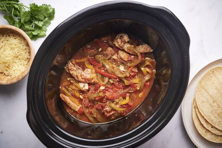 Slow cooker chicken fajitas with bell peppers, onions, sliced chicken breast, and tomatoes in a black slow crockpot with tortillas on the side.