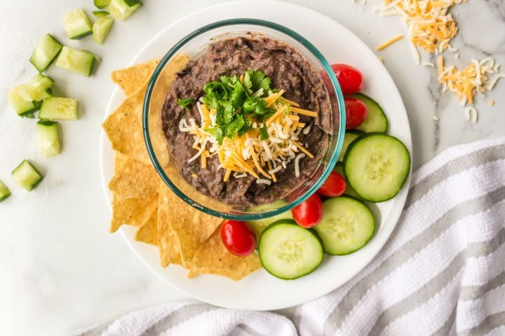 Easy black bean dip in a bowl with shredded cheese and cilantro with cucumbers, tomatoes, and chips on the side.