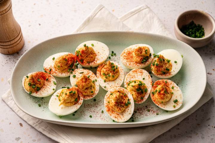 A plate of deviled eggs garnished with paprika and chives is set on a beige napkin. A small bowl of chives and a pepper mill are nearby.