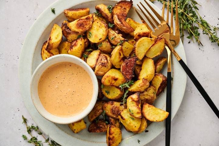 Easy roasted potatoes coated in olive oil and mustard on a white plate with dipping sauce and herbs.