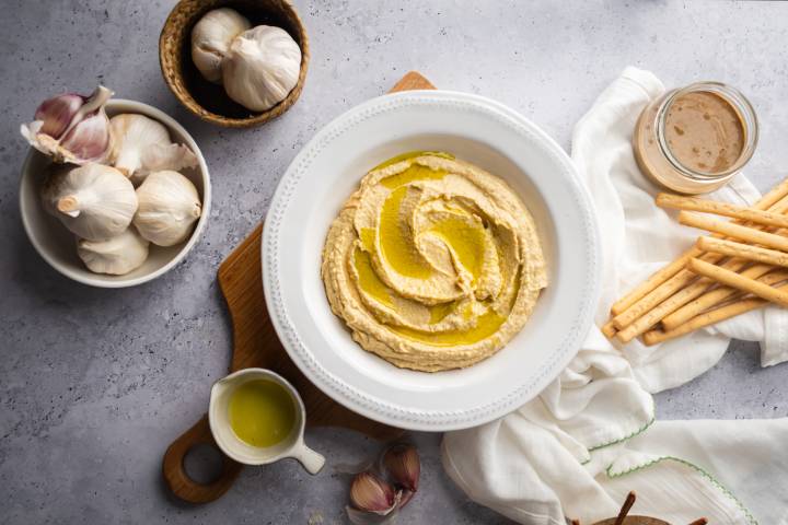 Garlic hummus swirled with olive oil with garlic cloves and breadsticks on the side.