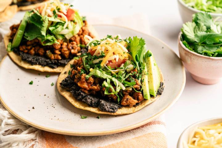 Loaded ground turkey tostadas topped with black beans, seasoned turkey, shredded lettuce, diced tomatoes, shredded cheese, fresh cilantro, and avocado slices, served on a plate with extra toppings nearby.