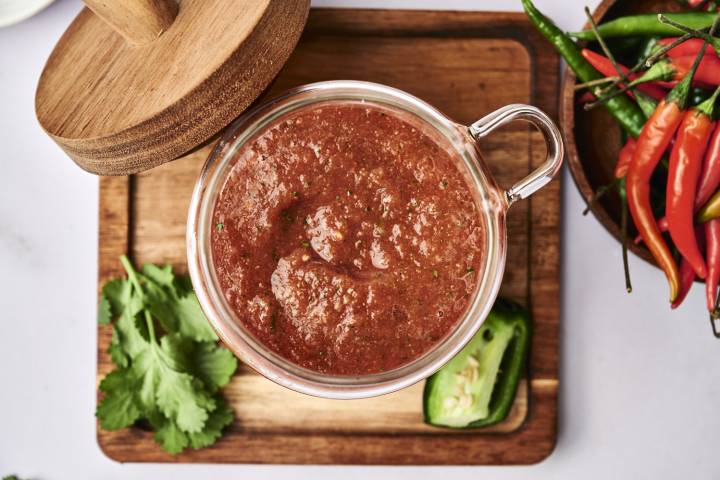 A glass jar filled with chunky salsa rests on a wooden board, surrounded by fresh cilantro and jalapeños, with vibrant chili peppers nearby.
