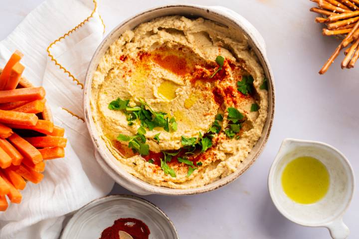 Hummus made with chickpeas in a bowl with olive oil, parsley, and paprika with carrots and pretzels on the side.